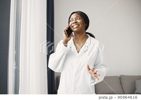 Young female black doctor standing in her office at clinic near coach. Woman wearing white medical coat. Brunette medical worker using a phone. Young female black doctor standing in her office at clinic near coach. Woman wearing white medical coat. Brunette medical worker using a phone. 90964616
