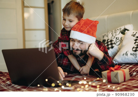 man i in santa hat with his little cute son laying with laptop on bed buying presents online man i in santa hat with his little cute son laying with laptop on bed buying presents online 90968230