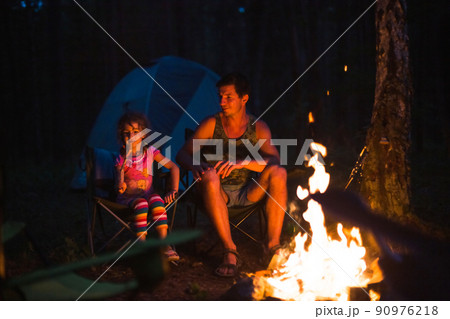 Dad and daughter sit at night by the fire in the open air in the summer in nature. Family camping trip, gatherings around the campfire. Father's Day, barbecue. Camping lantern and tent 90976218