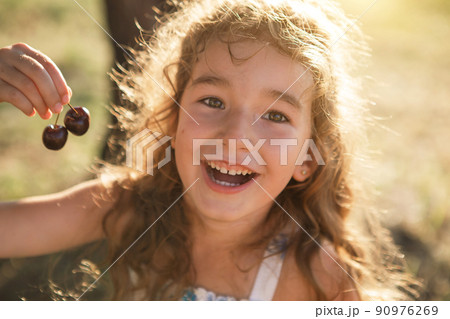 A cheerful sweet girl with cherry berries in her mouth. Funny summer portrait of a child with a cherry, gifts of summer, summer time. A cheerful sweet girl with cherry berries in her mouth. Funny summer portrait of a child with a cherry, gifts of summer, summer time. 90976269