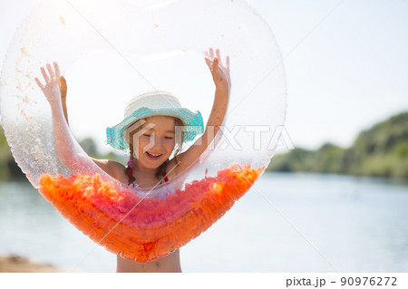 Girl in a hat stands on the river bank with a transparent inflatable circle in the shape of a heart with orange feathers inside. Beach holidays, swimming, tanning, sunscreens. Girl in a hat stands on the river bank with a transparent inflatable circle in the shape of a heart with orange feathers inside. Beach holidays, swimming, tanning, sunscreens. 90976272