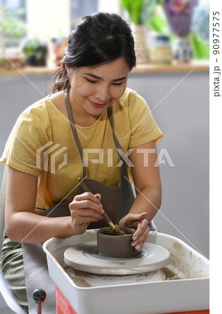 Smiling young woman in apron enjoy creating handmade potter in workshop. Indoors lifestyle activity, handicraft, hobbies concept 90977575