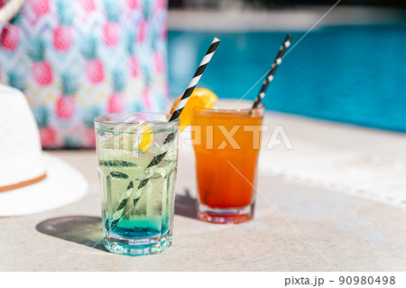 Tropical sparkling lemonade cocktails by the pool with pink beach bag and white hat in the background. Picture of glasses with orange and mint lemon fruit cocktails. Hello summer holiday vacation Tropical sparkling lemonade cocktails by the pool with pink beach bag and white hat in the background. Picture of glasses with orange and mint lemon fruit cocktails. Hello summer holiday vacation 90980498