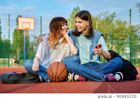 Student friends sitting on outdoor basketball court looking in smartphone 90983220