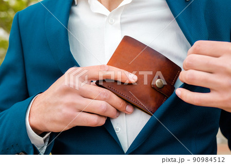 A businessman in a blue jacket and white shirt take off a brown leather wallet in a pocket. Close-up of the hands. The concept of style and fashion 90984512