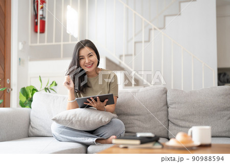 Beautiful attractive young Asian woman sitting on gray sofa and using digital tablet at living room in the morning. Beautiful attractive young Asian woman sitting on gray sofa and using digital tablet at living room in the morning. 90988594