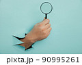 Male hand holds black magnifying glass in torn hole of blue background. Searching information concept 90995261