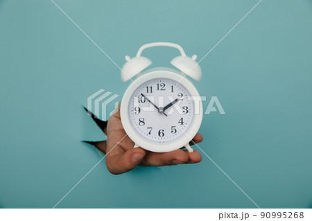 Hand holds alarm clock through a blue paper hole. Time management and deadline concept Hand holds alarm clock through a blue paper hole. Time management and deadline concept 90995268