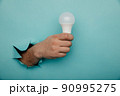 Hand holding an incandescent led light bulb from a torn hole in blue paper 90995275