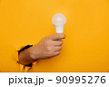 Hand holding an incandescent led light bulb from a torn hole in yellow paper 90995276