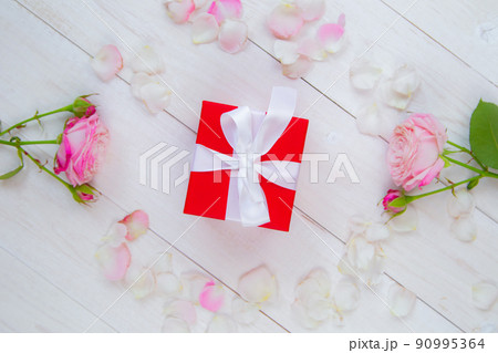 On a white wooden background there are roses and petals forming a circle, inside there is a gift in a red box and a white ribbon. High quality photo 90995364