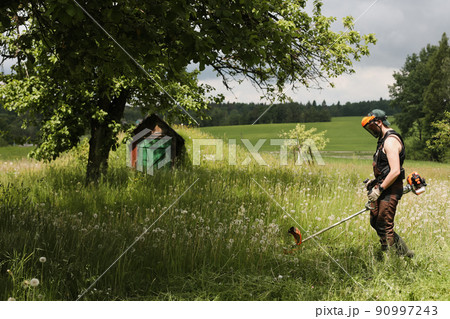 Man mowing tall grass with petrol lawn trimmer in the garden or backyard. Process of lawn trimming with hand mower Man mowing tall grass with petrol lawn trimmer in the garden or backyard. Process of lawn trimming with hand mower 90997243