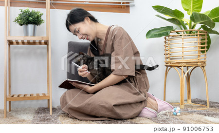 Asian woman sits happily writing a diary and playing with a cat in the living room, Beloved pet, Best friend pet, Happy area, Holidays at home, Relax and have fun, Sitting on the carpet with the cat. 91006753