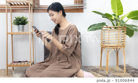 Young Asian woman happily using her mobile phone to chat or talking with her friends, Entertaining or relaxing using mobile phone technology, Stay at home for long weekend, Happy lifestyle. 91006844