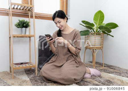 Young Asian woman happily using her mobile phone to chat or talking with her friends, Entertaining or relaxing using mobile phone technology, Stay at home for long weekend, Happy lifestyle. 91006845