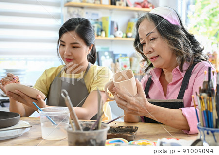 Woman painting a ceramic plate with acrylic colour with her grandmother in the workshop. 91009094