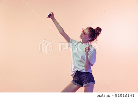 Portrait of young cheerful girl posing, taking selfie with mobile phone isolated over pink studio background 91013141