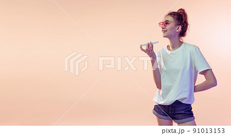 Portrait of young girl in white T-shirt recording voice message on phone, posing isolated over pink studio background in neon light 91013153