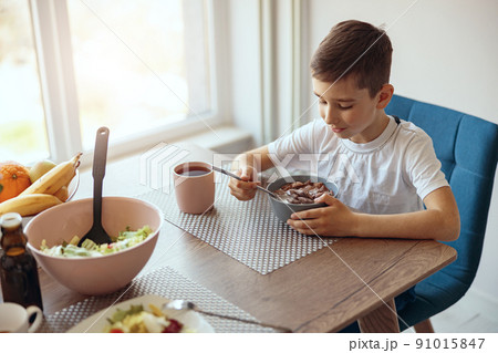 Caucasian little teen boy having cereals with milk and drinking tea at table in morning. Caucasian little teen boy having cereals with milk and drinking tea at table in morning. 91015847