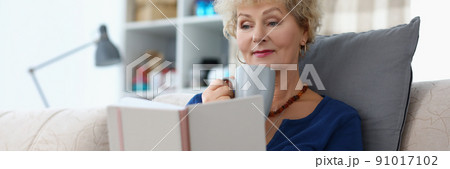 Old lady reading book and drinking cup of coffee being comfy on sofa at home 91017102