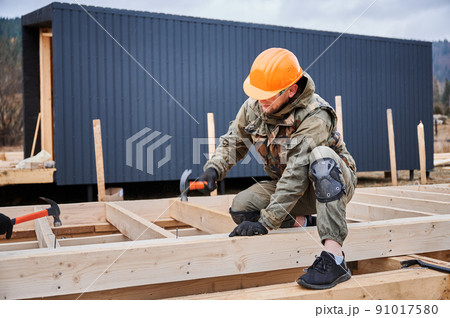 Man worker building wooden frame house on pile foundation. Carpenter hammering nail into wooden joist, using hammer. Carpentry concept. Man worker building wooden frame house on pile foundation. Carpenter hammering nail into wooden joist, using hammer. Carpentry concept. 91017580