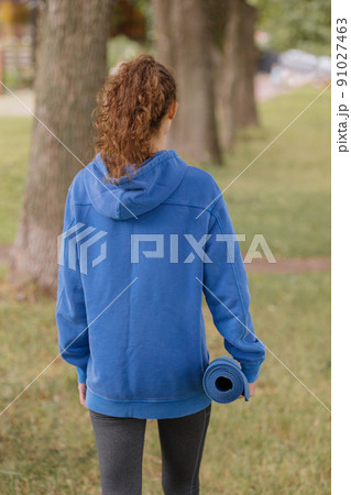 a European woman in a blue hoodie with a blue yoga mat chooses a place to do sports and meditation in the park. 91027463