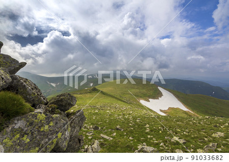 Wide panorama of lit by sun mountain plateau with green grass, patches of snow and big boulders on distant mountains under cloudy sky background. Beauty of nature, tourism and traveling concept. 91033682