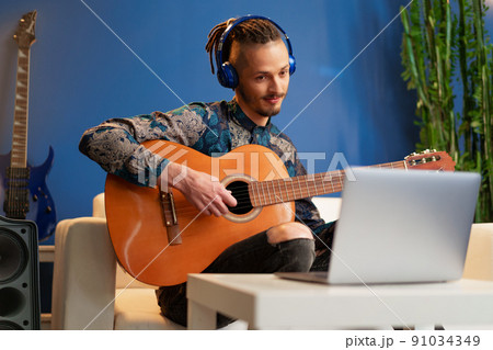 Young man sitting in his room and watching guitar tutorial on laptop 91034349