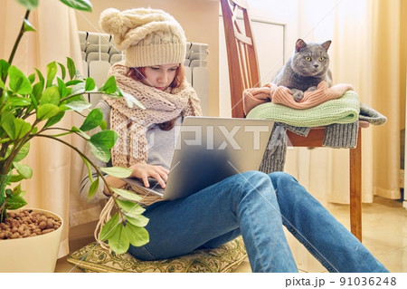 Preteen girl in scarf hat using laptop, warming near heating radiator with pet gray cat 91036248