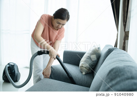 Asian young woman using vacuum machine to cleanup the dust on curtain in living room close up. Housework and chores in daily life concept. Housekeeper cleaning a white curtain. 91040980
