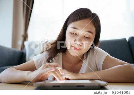 Happy Asian young woman relaxing in the living room and watching a contents and chatting with her friends and drawing an illustration on tablet. Woman enjoy watching video and drawing on tablet. 91041005