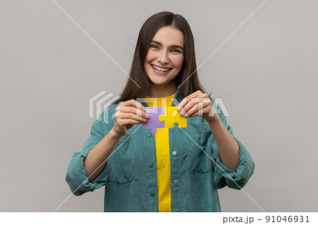 Smiling happy woman holding colorful pieces of puzzle, two parts of one, symbol of connection and union, association, wearing casual style jacket. Indoor studio shot isolated on gray background. Smiling happy woman holding colorful pieces of puzzle, two parts of one, symbol of connection and union, association, wearing casual style jacket. Indoor studio shot isolated on gray background. 91046931