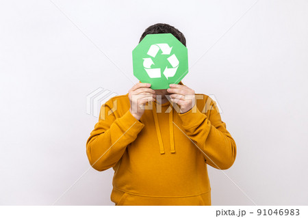 Anonymous male person hiding face behind green recycling sign, garbage sorting and environment protection, wearing urban style hoodie. Indoor studio shot isolated on white background. 91046983