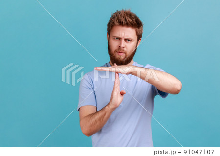 I need more time. Portrait of bearded man showing time out gesture, looking with imploring eyes, hurry to meet deadline, begging for more time. Indoor studio shot isolated on blue background. 91047107