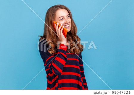 Portrait of positive happy woman wearing striped casual style sweater, having pleasant conversation, standing and talking phone with smile.Indoor studio shot isolated on blue background. 91047221