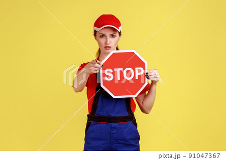 Portrait of serious worker woman showing holding red stop road sign, pointing at camera, prohibitions and restrictions, wearing overalls and red cap. Indoor studio shot isolated on yellow background. Portrait of serious worker woman showing holding red stop road sign, pointing at camera, prohibitions and restrictions, wearing overalls and red cap. Indoor studio shot isolated on yellow background. 91047367
