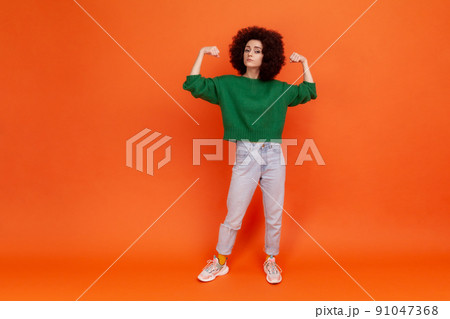 Full length portrait of woman with Afro hairstyle in green sweater raising arms showing biceps as metaphor of female power and independence, feminism. Indoor studio shot isolated on orange background. Full length portrait of woman with Afro hairstyle in green sweater raising arms showing biceps as metaphor of female power and independence, feminism. Indoor studio shot isolated on orange background. 91047368