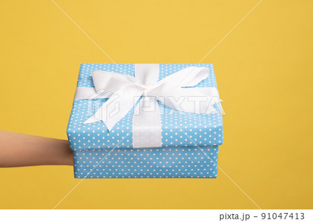 Closeup side view of woman hand holding out blue gift box with ribbon, giving present on holiday, bonuses and surprises concept. Indoor studio shot isolated on yellow background. 91047413