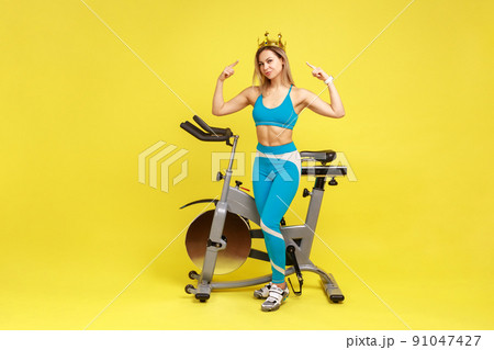Full length portrait of woman sport coach standing pointing at golden crown on her head, being queen of fitness, wearing blue sportswear. Indoor studio shot isolated on yellow background. Full length portrait of woman sport coach standing pointing at golden crown on her head, being queen of fitness, wearing blue sportswear. Indoor studio shot isolated on yellow background. 91047427