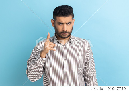 Portrait of serious strict bearded businessman standing with admonishing gesture, scolding for mistake and warning, wearing striped shirt. Indoor studio shot isolated on blue background. 91047750