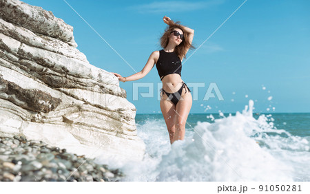 a girl in a swimsuit poses in the spray of waves at a white rock on the seashore a girl in a swimsuit poses in the spray of waves at a white rock on the seashore 91050281
