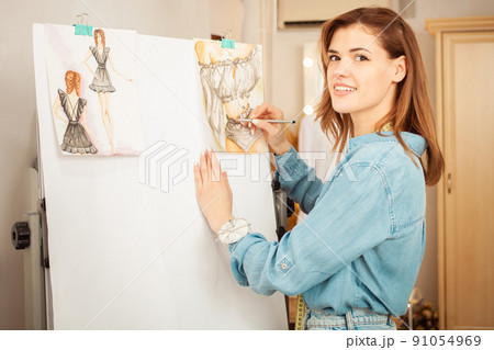 Portrait of beautiful seamstress woman. Fashionable sewing studio indoors. Blue shirt, brown hair, cozy interior. Draws clothes 91054969