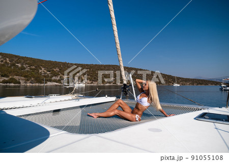 Beautiful young blond woman in bikini sitting on catamaran bow at sunny summer day 91055108