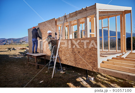 Carpenters mounting wooden OSB panel on the wall of future cottage. Men workers building wooden frame house on pile foundation in the Scandinavian style barnhouse. Carpentry and construction concept. Carpenters mounting wooden OSB panel on the wall of future cottage. Men workers building wooden frame house on pile foundation in the Scandinavian style barnhouse. Carpentry and construction concept. 91057100