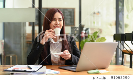 Thoughtful asian woman in business suit drinking hot coffee and using laptop at modern coffee shop 91062590