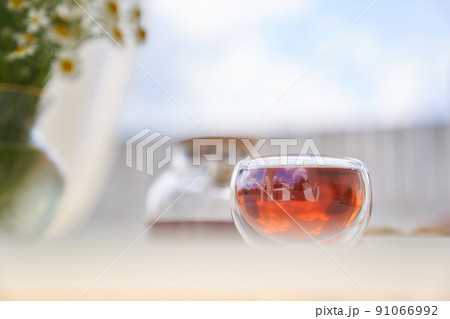 Transparent glass Cup of tea in nature. The concept of breakfast in the backyard of the house. Early morning, tea and kettle. out of focus, chamomile flowers in the background of blue sky. 91066992