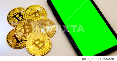 Bitcoin coins in golden metal with a smarthphone beside it, green screen area copy space 91068020