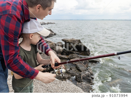 Father and son fishing. Dad shows his son how to hold the spinning and spin the reel. Fishing training on a pond or river. Caring parent concept. Father and son fishing. Dad shows his son how to hold the spinning and spin the reel. Fishing training on a pond or river. Caring parent concept. 91073994