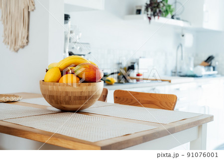 Home kitchen counter table with selective focus on bamboo bowl with exotic fruits on it with blurred background of modern cozy white kitchen. Home interior design details. Copy space. Home kitchen counter table with selective focus on bamboo bowl with exotic fruits on it with blurred background of modern cozy white kitchen. Home interior design details. Copy space. 91076001