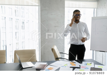 Smiling African businessman talks on cellphone standing in modern workplace Smiling African businessman talks on cellphone standing in modern workplace 91078869